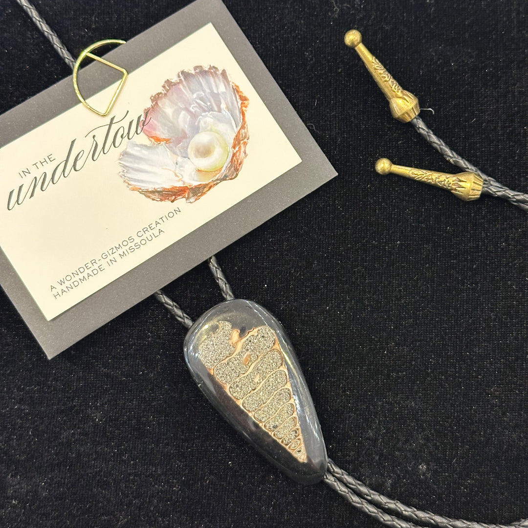 A cross section of a fossilized shell as the centerpiece on  bolo tie with a black cord and metal tips made in Missoula by IN the Undertow