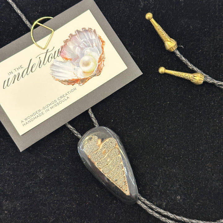 A cross section of a fossilized shell as the centerpiece on  bolo tie with a black cord and metal tips made in Missoula by IN the Undertow