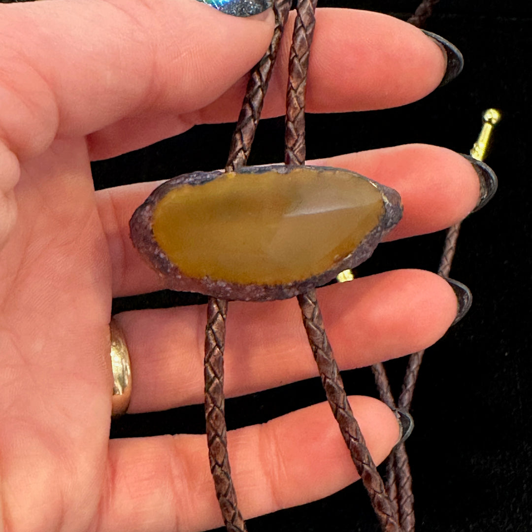 A close up of a hand holding a polished agate bolo tie on a leather cord