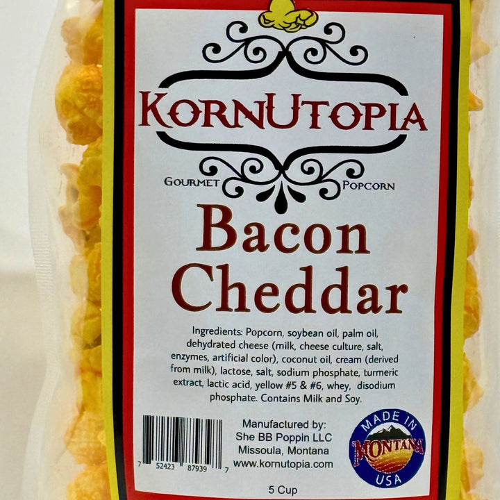 A close-up of a packaged snack bag labeled KornUtopia Gourmet Popcorn, Bacon Cheddar flavor. The front label is white with red and black text, and yellow popcorn is visible through the clear plastic packaging on both sides. Mae in Montana USA