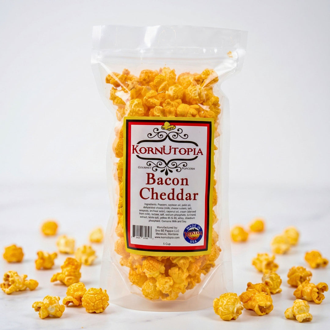 A clear plastic bag of Korn Utopia Bacon Cheddar popcorn standing upright on a white background, with scattered golden-yellow popcorn kernels around the base.