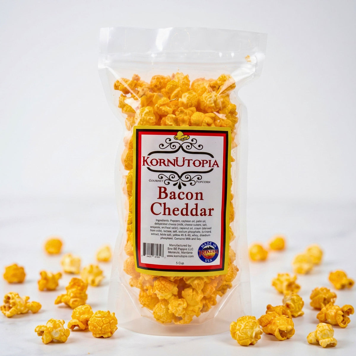 A clear plastic bag of Korn Utopia Bacon Cheddar popcorn standing upright on a white background, with scattered golden-yellow popcorn kernels around the base.