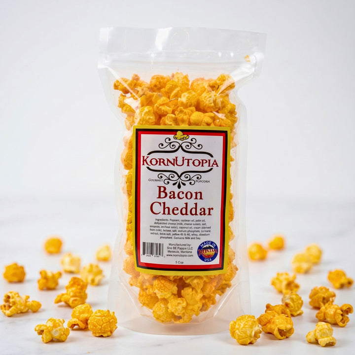 A clear plastic bag of Korn Utopia Bacon Cheddar popcorn standing upright on a white background, with scattered golden-yellow popcorn kernels around the base.