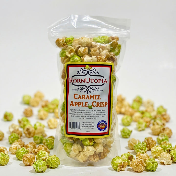 A clear plastic bag of KornUtopia Caramel Apple Crisp popcorn standing upright on a white surface, with scattered caramel-apple popcorn pieces around it. The front label shows the KornUtopia brand and the flavor name.