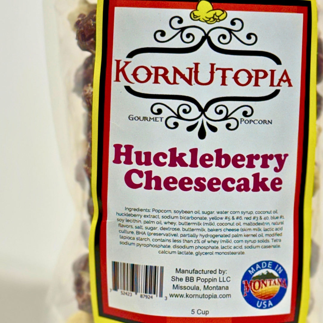 A close-up of a Kornutopia Gourmet Popcorn package labeled Huckleberry Cheesecake, showing the front label with red and black text on a white background and a yellow border. Made in Montana USA