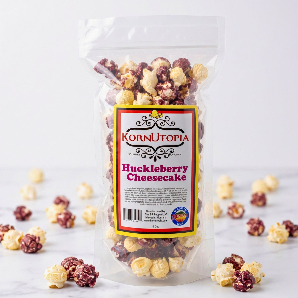 A clear bag of KornUtopia Huckleberry Cheesecake Gourmet Popcorn on a white marble surface with purple and white popcorn scattered around the base.