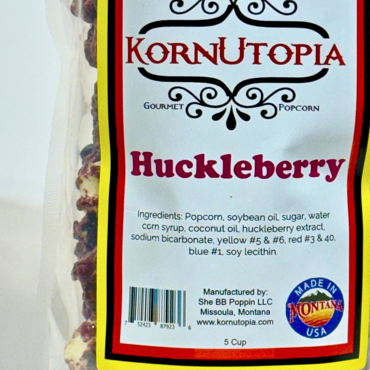 A close-up of a KornUtopia Gourmet Huckleberry popcorn bag, showing the white label with red and black text, a barcode, and a &