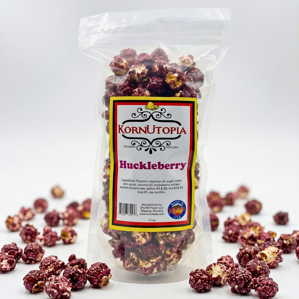 A clear plastic bag of KornUtopia Huckleberry popcorn with a white, red, and yellow label, shown on a white background with scattered huckleberry popcorn kernels around the base.