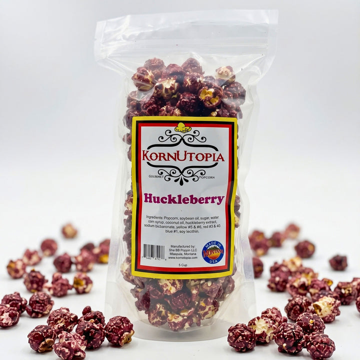 A clear plastic bag of KornUtopia Huckleberry popcorn with a white, red, and yellow label, shown on a white background with scattered huckleberry popcorn kernels around the base.