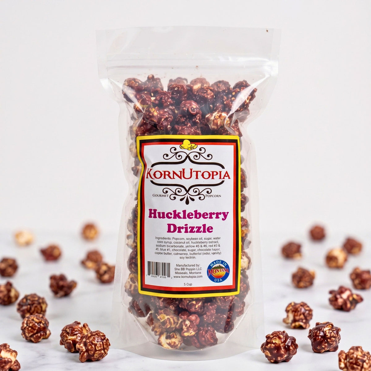 A clear stand-up pouch of Kornutoпия Huckleberry Drizzle popcorn, filled with glossy brown and reddish-brown coated popcorn kernels, with a few scattered around the base on a white surface.