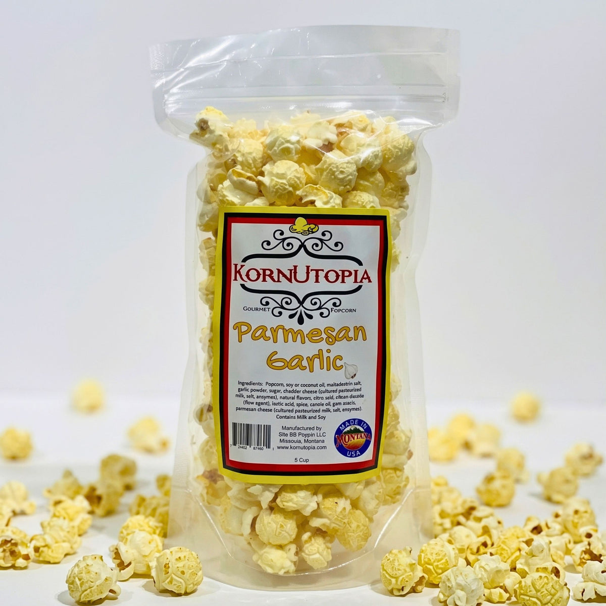 A clear plastic bag of KornUtopia Parmesan Garlic popcorn standing upright on a white surface, with scattered popcorn kernels around the base and a white studio background.