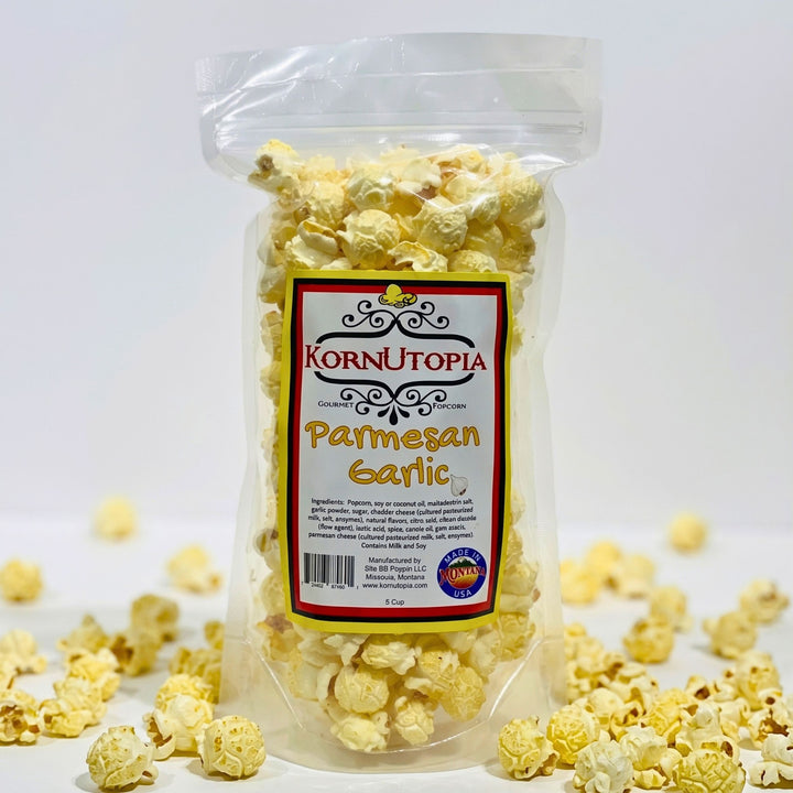 A clear plastic bag of KornUtopia Parmesan Garlic popcorn standing upright on a white surface, with scattered popcorn kernels around the base and a white studio background.