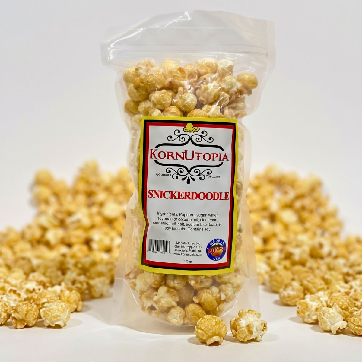 A clear plastic bag of KornUtopia Snickerdoodle popcorn, showing the golden-brown caramelized popcorn inside, with more popcorn scattered around it on a white background.