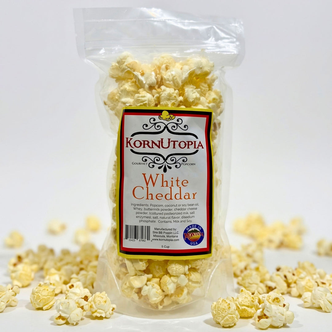 A clear plastic bag of Kornutopia White Cheddar popcorn with a white, red, and black label, set against a white background with scattered popcorn kernels around the base.