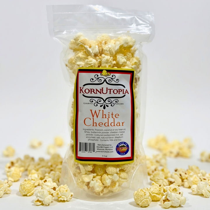 A clear plastic bag of Kornutopia White Cheddar popcorn with a white, red, and black label, set against a white background with scattered popcorn kernels around the base.