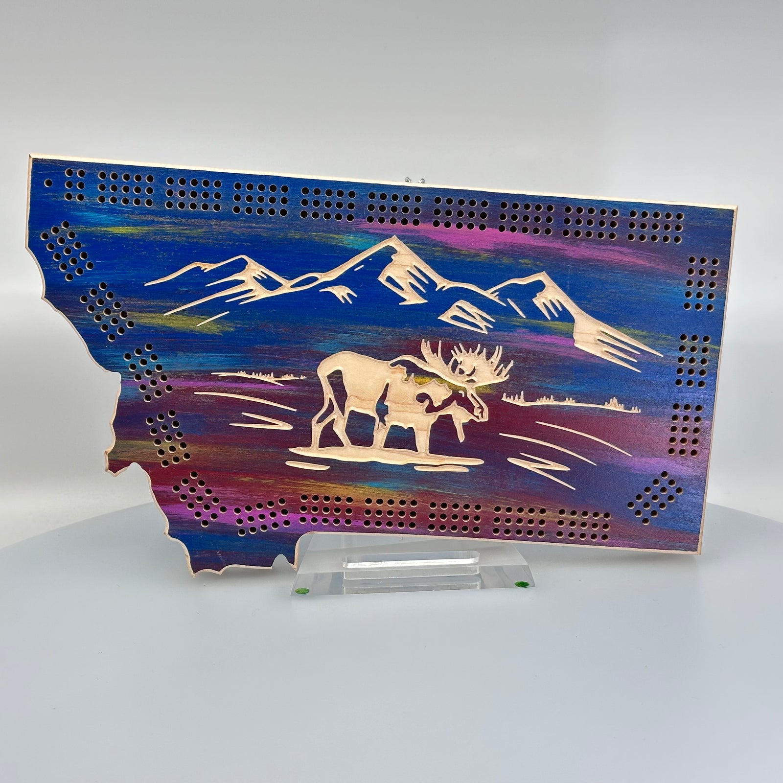 Knotty Pine Woodworks HandmadeMontana Moose Cribbage Board Sunset (front)--this made in Montana wooden cribbage board is shaped like the outline of the state of Montana, painted in sunset tones, and features an engraved moose standing in front of mountain peaks on the front.