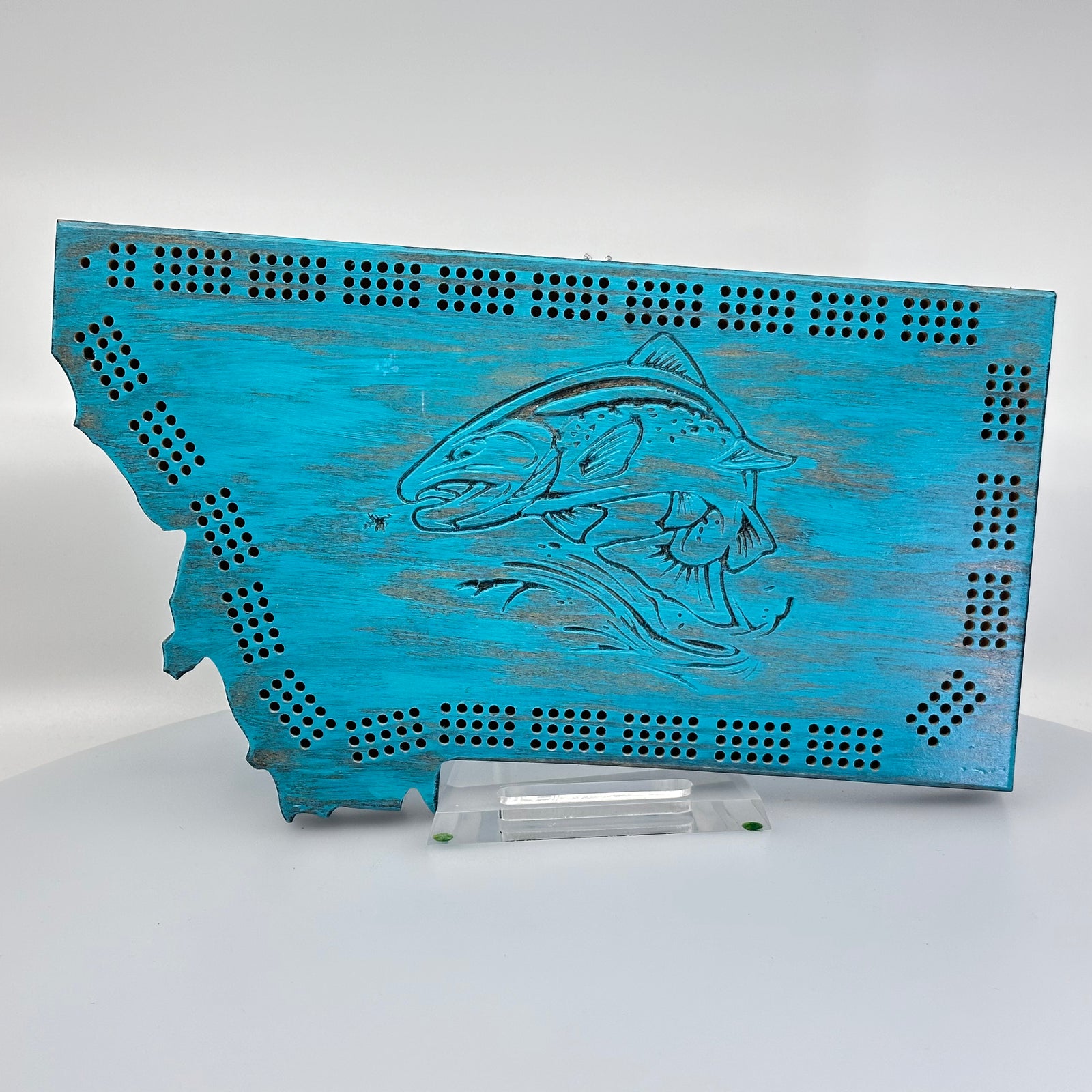 Knotty Pine Woodworks Handmade Montana Trout Cribbage Board Turquoise (front)--this made in Montana wooden cribbage board is shaped like the outline of the state of Montana, in weathered turquoise tones, and features an engraved trout jumping out of water on the front.