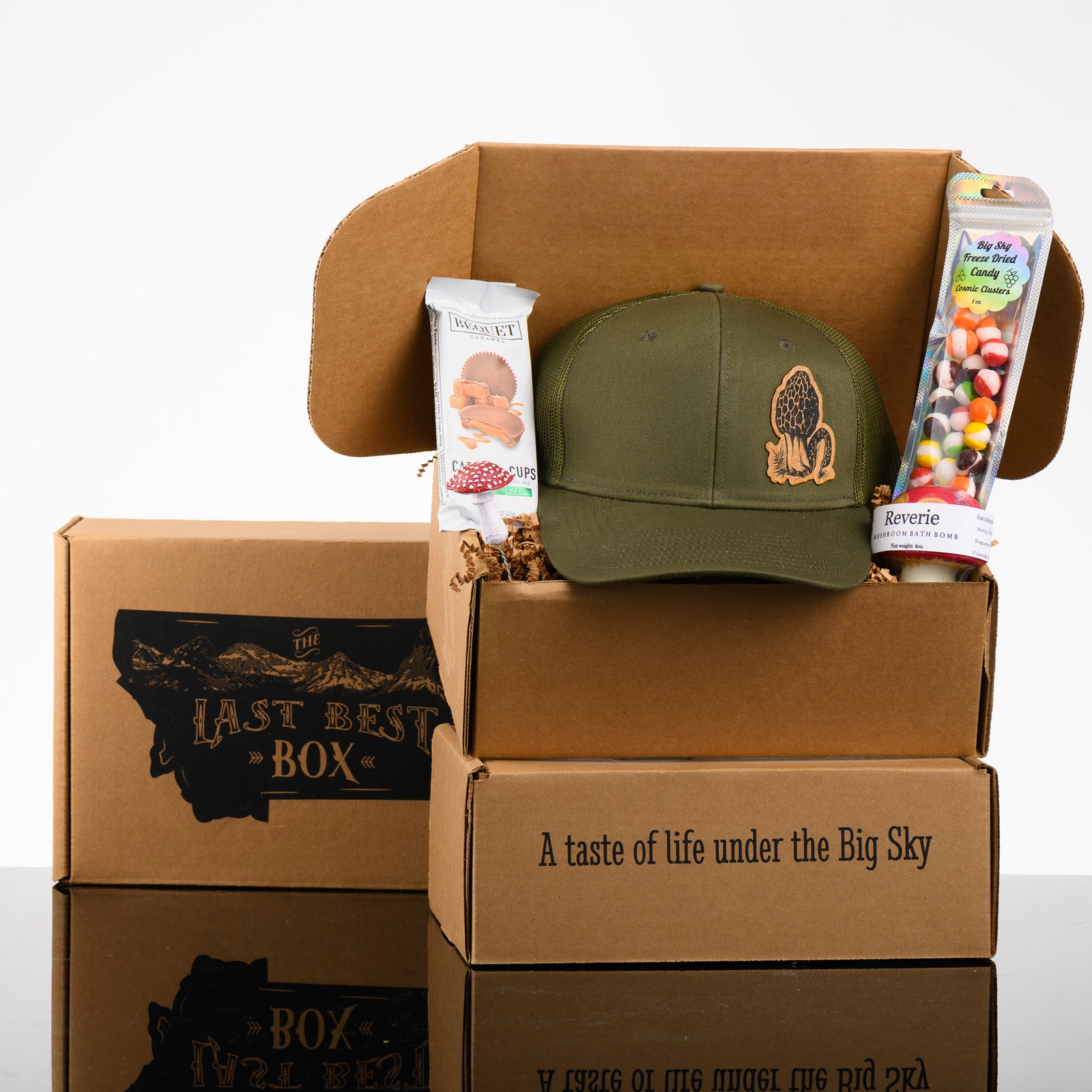 A stack of Montana made gift boxes with a green hat with a morel mushroom wood patch, a mushroom bath bomb, freeze dried skittles, a mushroom plant decoration, and caramel chocolate cups.
