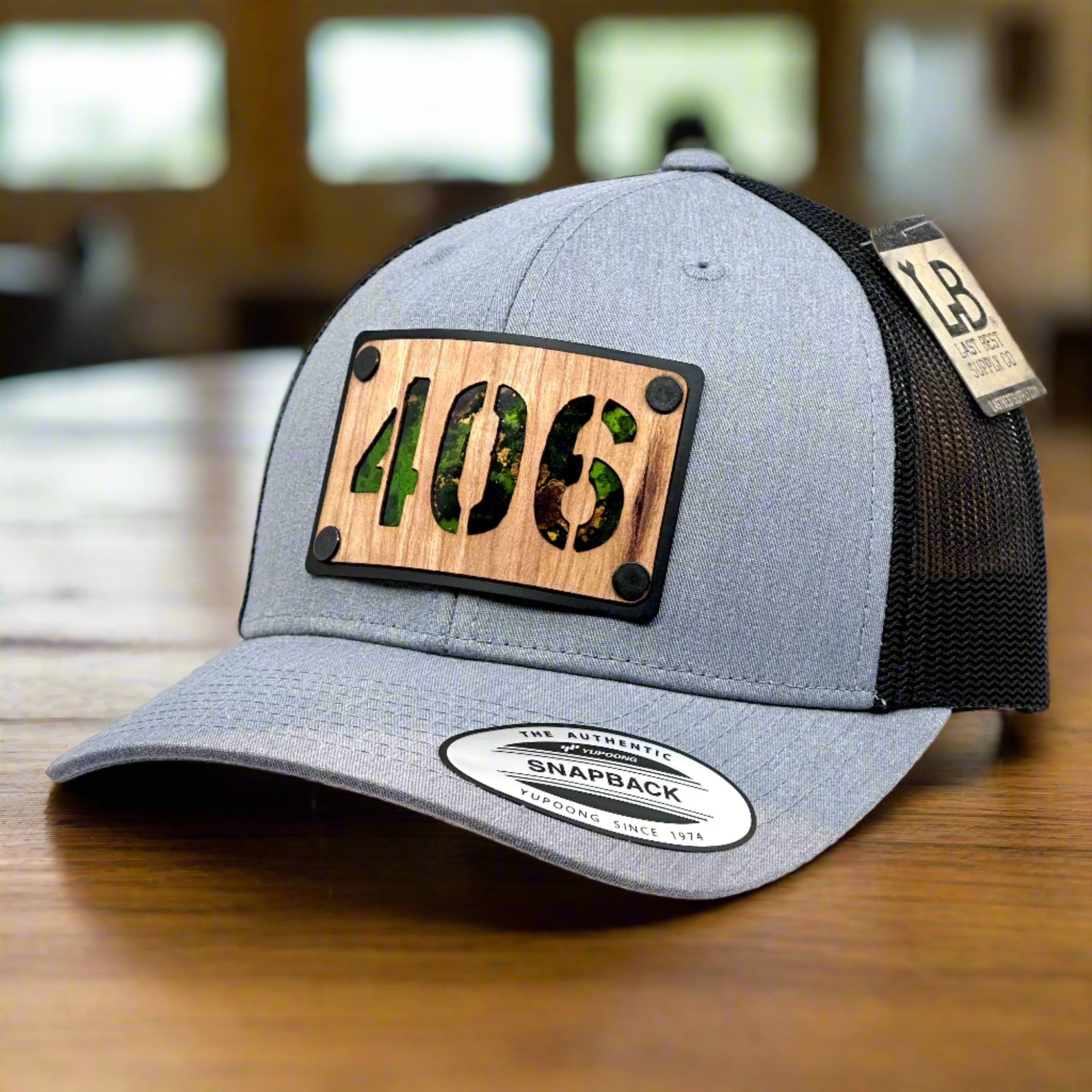 3/4 view of a 406 Cherry Wood & Green Copper Plate Patch Flexfit Trucker Hat featuring a rectangular cherry wood patch with the Montana area code 406 cut out, over an anodized aluminum metal base plate patinated green copper, and metal rivets—handmade in Montana, USA by Last Best Supply Co.