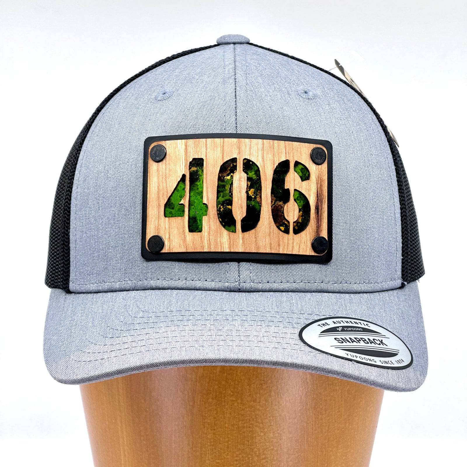 Front of a 406 Cherry Wood & Green Copper Plate Patch Flexfit Trucker Hat featuring a rectangular cherry wood patch with the Montana area code 406 cut out, over an anodized aluminum metal base plate patinated green copper, and metal rivets—handmade in Montana, USA by Last Best Supply Co.