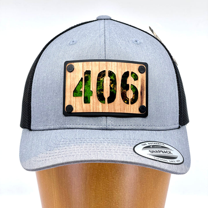 Front of a 406 Cherry Wood & Green Copper Plate Patch Flexfit Trucker Hat featuring a rectangular cherry wood patch with the Montana area code 406 cut out, over an anodized aluminum metal base plate patinated green copper, and metal rivets—handmade in Montana, USA by Last Best Supply Co.