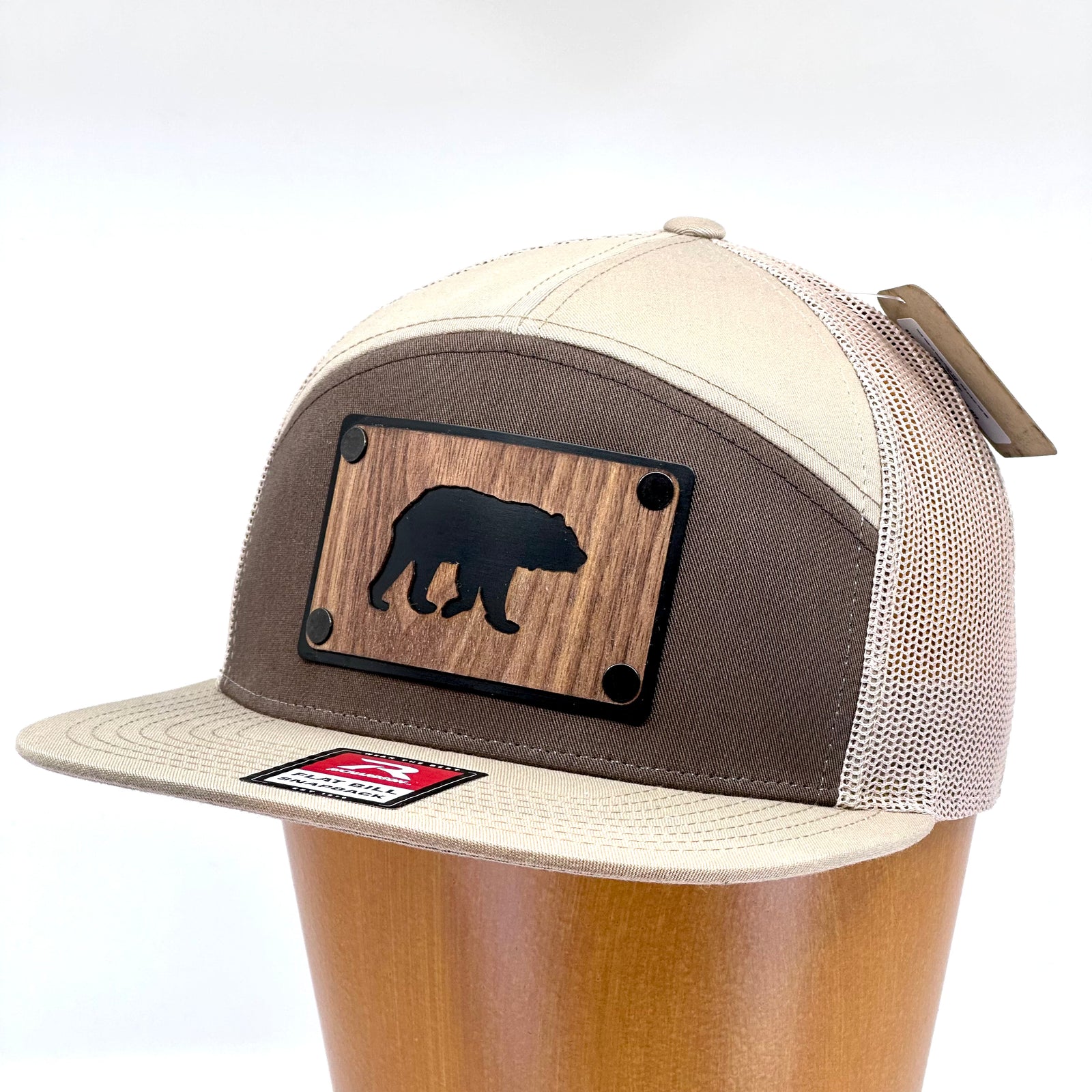 3/4 view of a Bear Walnut Wood Patch Hat Flat Bill Hat featuring a rectangular walnut wood patch with a bear silhouette cut out, over a black aluminum metal base plate, and metal rivets, attached to a pale khaki, brown, and white Richardson 7-panel trucker hat—handmade in Montana, USA by Last Best Supply Co.