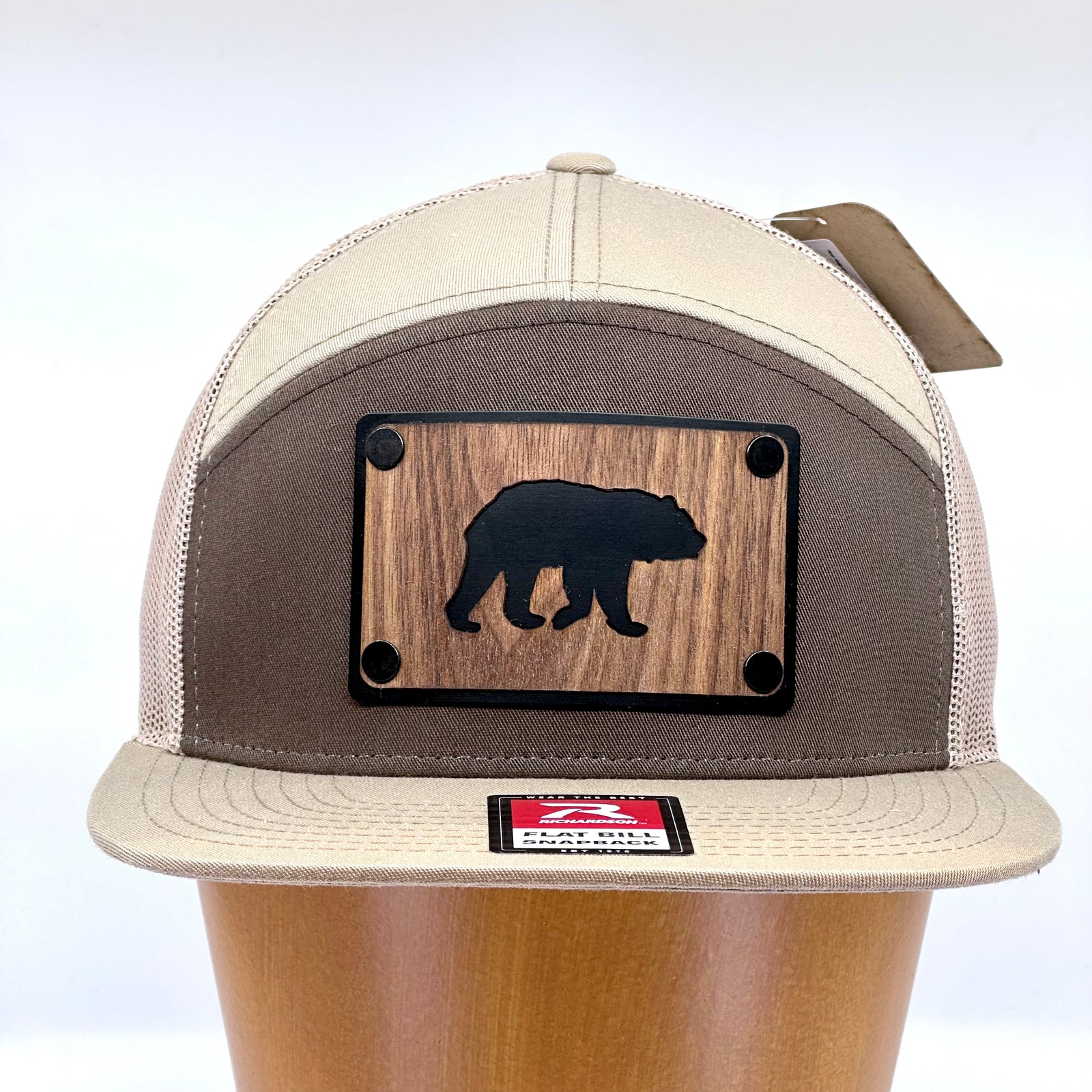 Front of a Bear Walnut Wood Patch Hat Flat Bill Hat featuring a rectangular walnut wood patch with a bear silhouette cut out, over a black aluminum metal base plate, and metal rivets, attached to a pale khaki, brown, and white Richardson 7-panel trucker hat—handmade in Montana, USA by Last Best Supply Co.