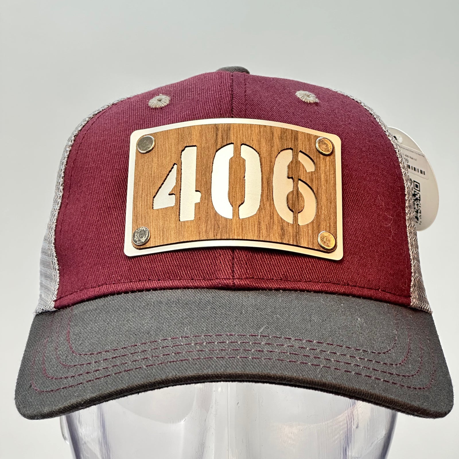 The front of a 406 Walnut Wood & Metal Patch Maroon, Charcoal and Grey Trucker Hat, featuring a rectangular, real cherry wood veneer patch with a precision-cut “406” cutout, laminated to a silver anodized metal plate, and then riveted to a maroon and charcoal grey mesh trucker hat—handmade in Montana, USA by Last Best Supply Co.