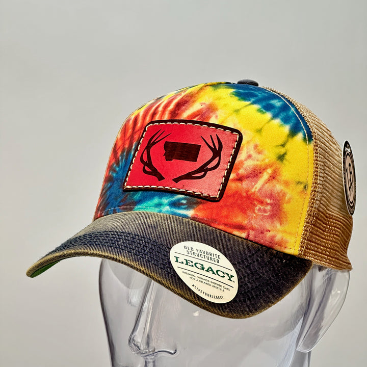 3/4 view of a Antlers Montana Tie Dye Premium Leather Patch Trucker Hat, featuring a rectangular real hand-dyed leather patch etched with a silhouette of Montana nestled between a pair of elk antlers, securely stitched with thick thread onto the structured front of a premium, rainbow tie-dyed trucker hat—handmade in Montana, USA by Last Best Supply Co.