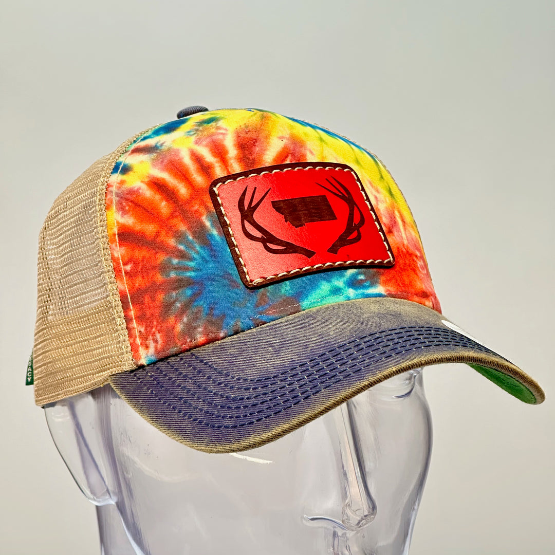 3/4 view of an Antlers Montana Tie Dye Premium Leather Patch Trucker Hat, featuring a rectangular real hand-dyed leather patch etched with a silhouette of Montana nestled between a pair of elk antlers, securely stitched with thick thread onto the structured front of a premium, rainbow tie-dyed trucker hat—handmade in Montana, USA by Last Best Supply Co.