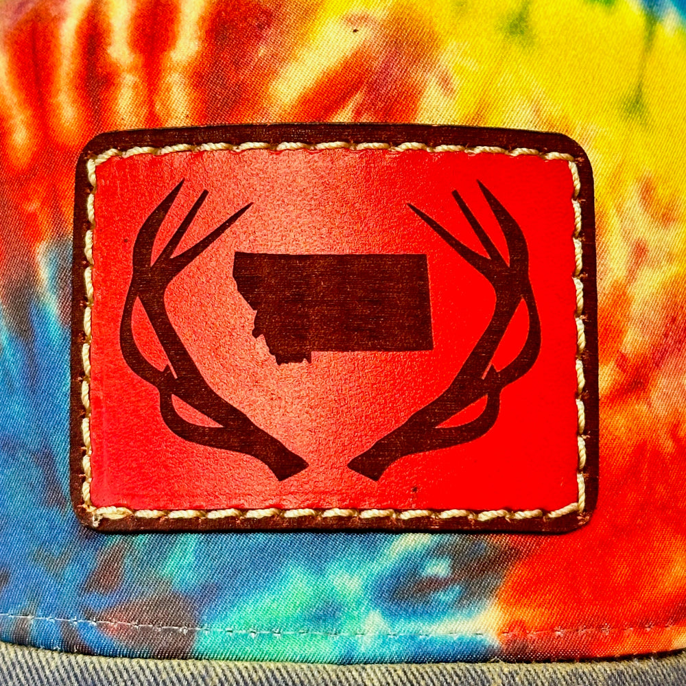 Patch detail of an Antlers Montana Tie Dye Premium Leather Patch Trucker Hat, featuring a rectangular real hand-dyed leather patch etched with a silhouette of Montana nestled between a pair of elk antlers, securely stitched with thick thread onto the structured front of a premium, rainbow tie-dyed trucker hat—handmade in Montana, USA by Last Best Supply Co.
