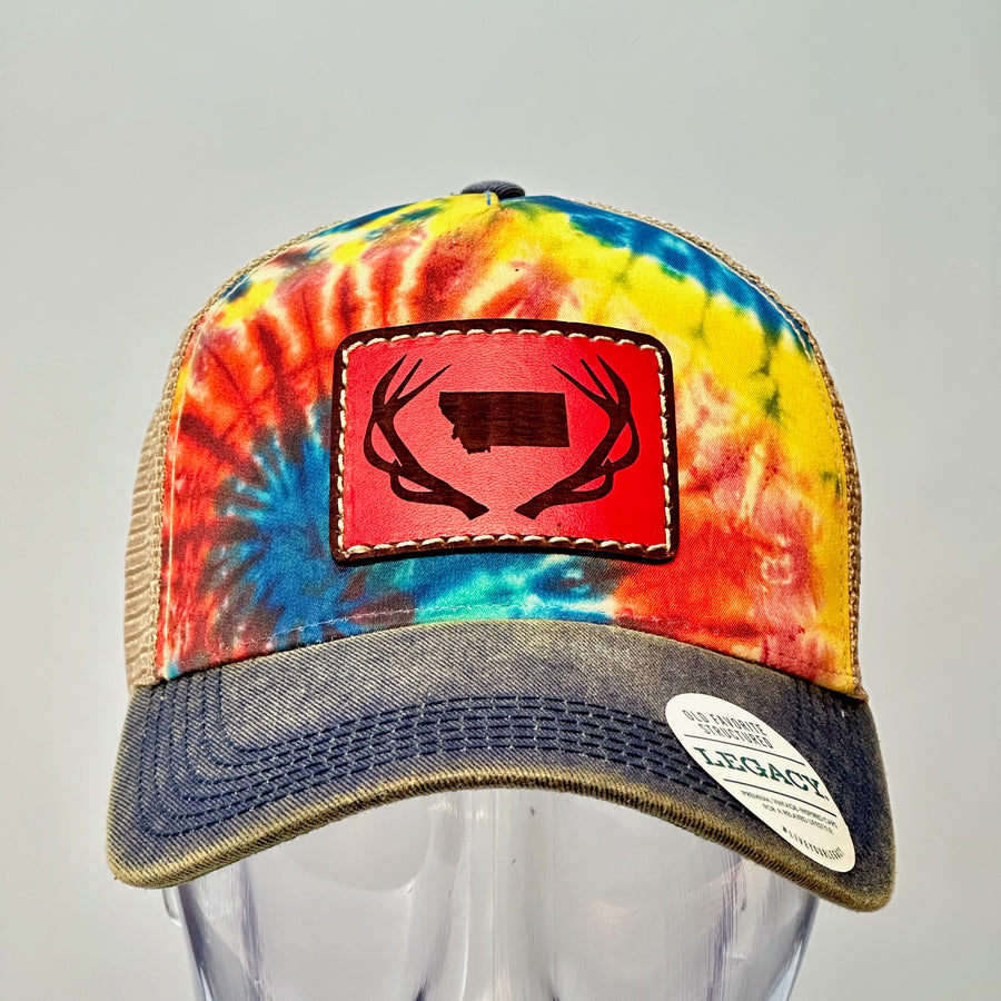 Front of an Antlers Montana Tie Dye Premium Leather Patch Trucker Hat, featuring a rectangular real hand-dyed leather patch etched with a silhouette of Montana nestled between a pair of elk antlers, securely stitched with thick thread onto the structured front of a premium, rainbow tie-dyed trucker hat—handmade in Montana, USA by Last Best Supply Co.