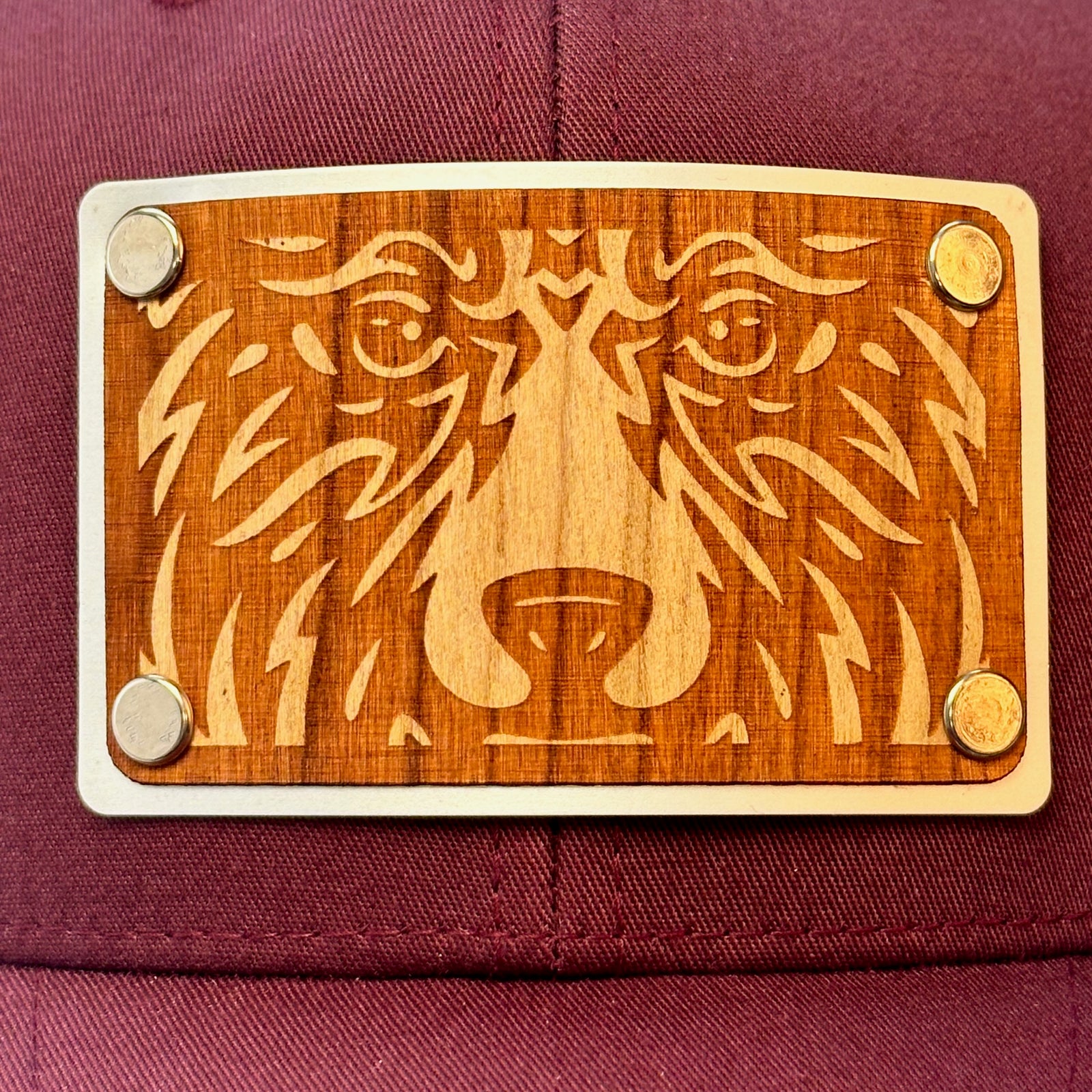 Patch detail of a Bear Face Cherry Wood & Silver Metal Patch Maroon and Grey Trucker Hat, featuring a rectangular, real cherry wood veneer patch etched with a bold bear face design, laminated to a silver anodized metal plate, and then riveted to a maroon and charcoal grey mesh trucker hat—handmade in Montana, USA by Last Best Supply Co.
