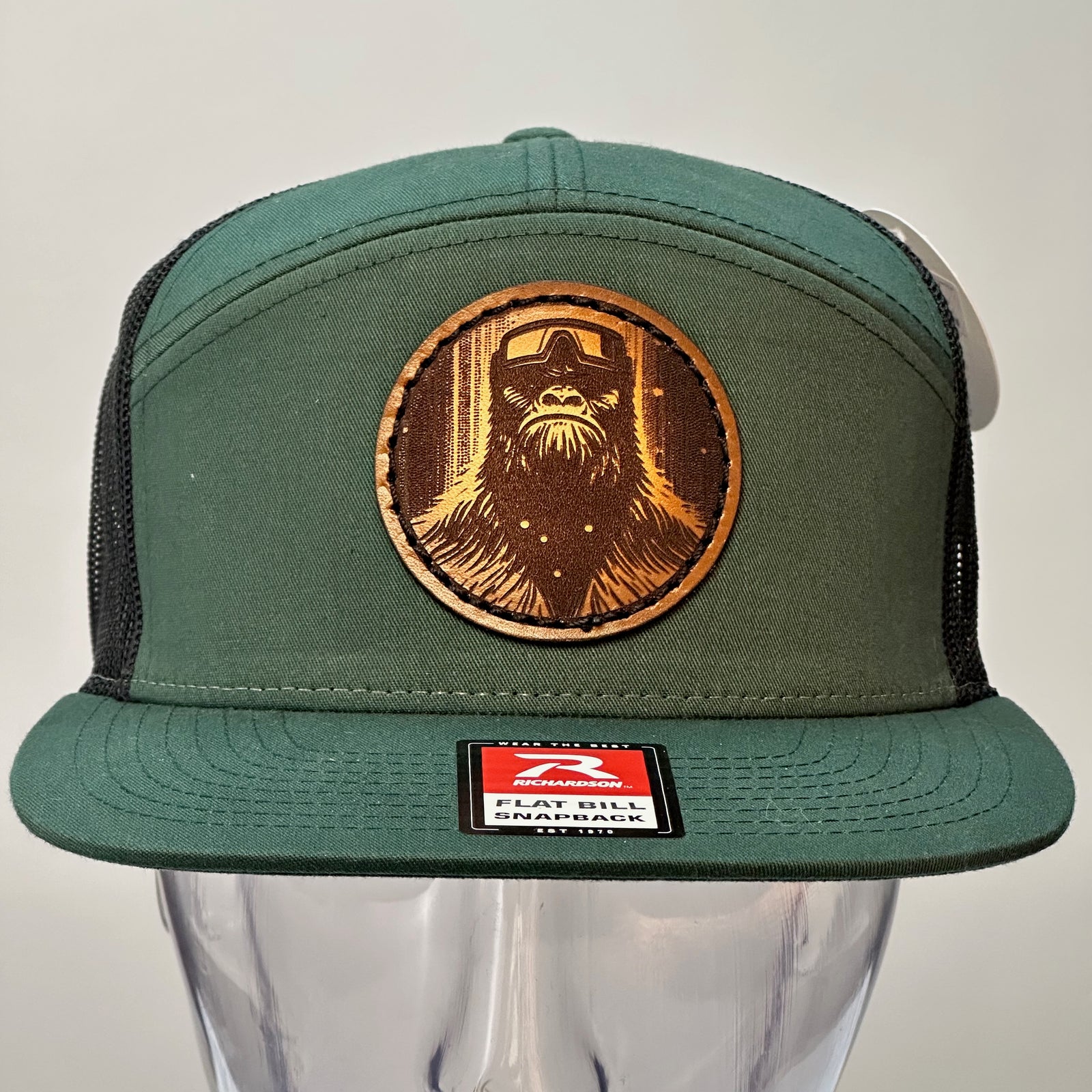 The front of a Bigfoot Face Premium Leather Patch Forest Green Flat Bill Hat, featuring a circular hand-dyed leather patch etched with Bigfoot’s face, securely stitched with thick thread onto the structured front of a green and black Richardson flat bill trucker hat—handmade in Montana, USA by Last Best Supply Co.
