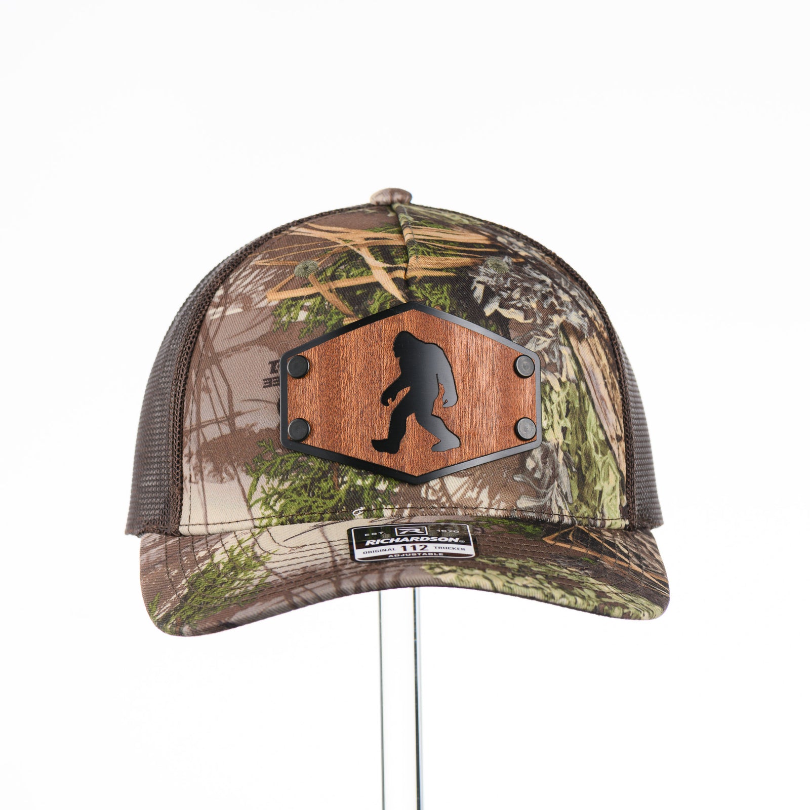 The front of a Bigfoot Mahogany Wood Hex Patch Camo Trucker Hat, featuring a hexagonal real mahogany wood veneer patch with a cutout silhouette of Bigfoot, laminated to a black anodized metal plate, and riveted to a camo and brown Richardson 112 trucker hat—handmade in Montana, USA by Last Best Supply Co.