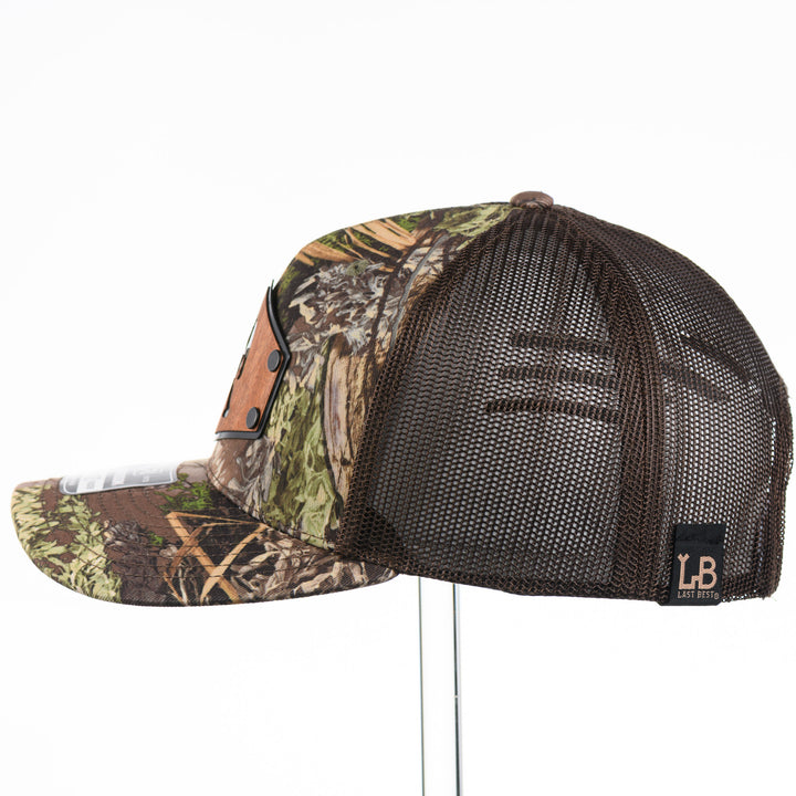 Side of a Bigfoot Mahogany Wood Hex Patch Camo Trucker Hat, featuring a hexagonal real mahogany wood veneer patch with a cutout silhouette of Bigfoot, laminated to a black anodized metal plate, and riveted to a camo and brown Richardson 112 trucker hat—handmade in Montana, USA by Last Best Supply Co.