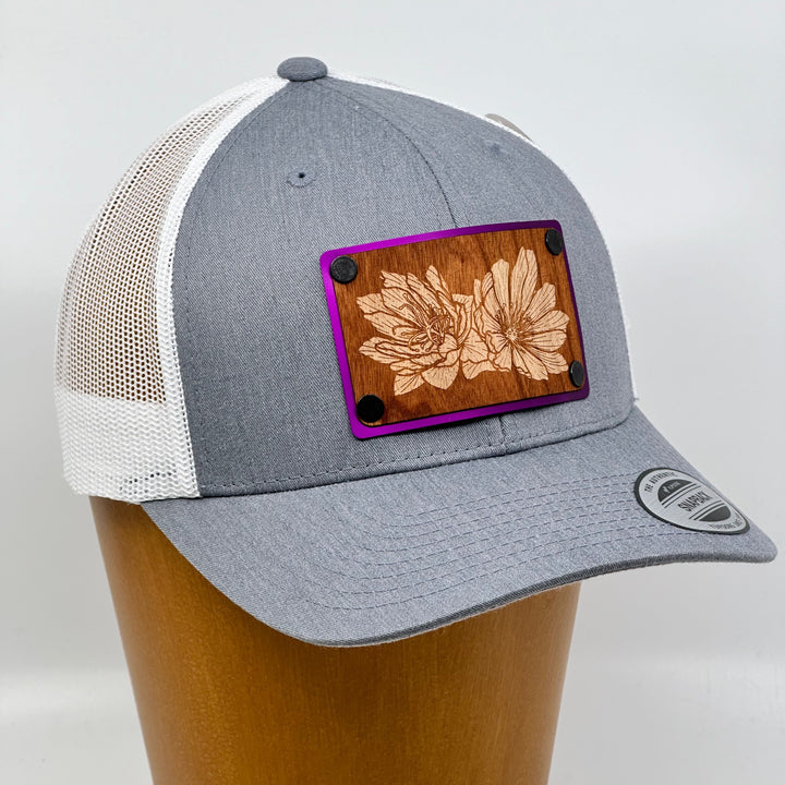 3/4 view of a Bitterroot Cherry Wood Patch Grey & White Trucker Hat, featuring a rectangular real cherry wood veneer patch with an intricate etched bitterroot flower design, laminated to a magenta anodized metal plate, and riveted to a grey and white mesh trucker hat—handmade in Montana, USA by Last Best Supply Co.