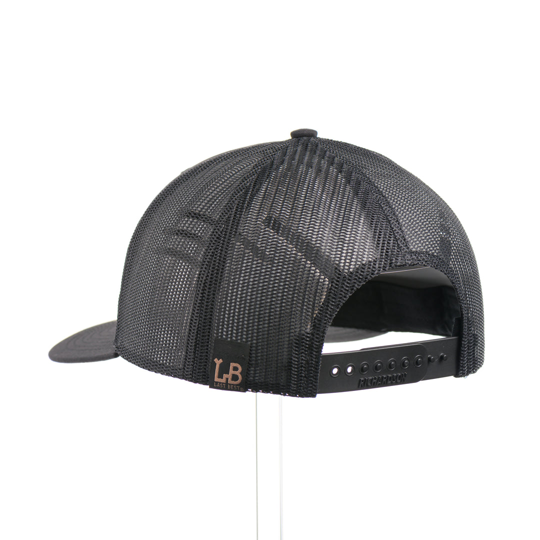 The back of a Black Grizzly Bear Teal Copper & Mahogany Wood Plate Patch Trucker Hat, featuring a rectangular real mahogany wood veneer patch with cut-out bear silhouette and teal copper inlay, laminated to a black anodized metal plate, and riveted to a black Richardson 112 trucker hat—handmade in Montana, USA by Last Best Supply Co.