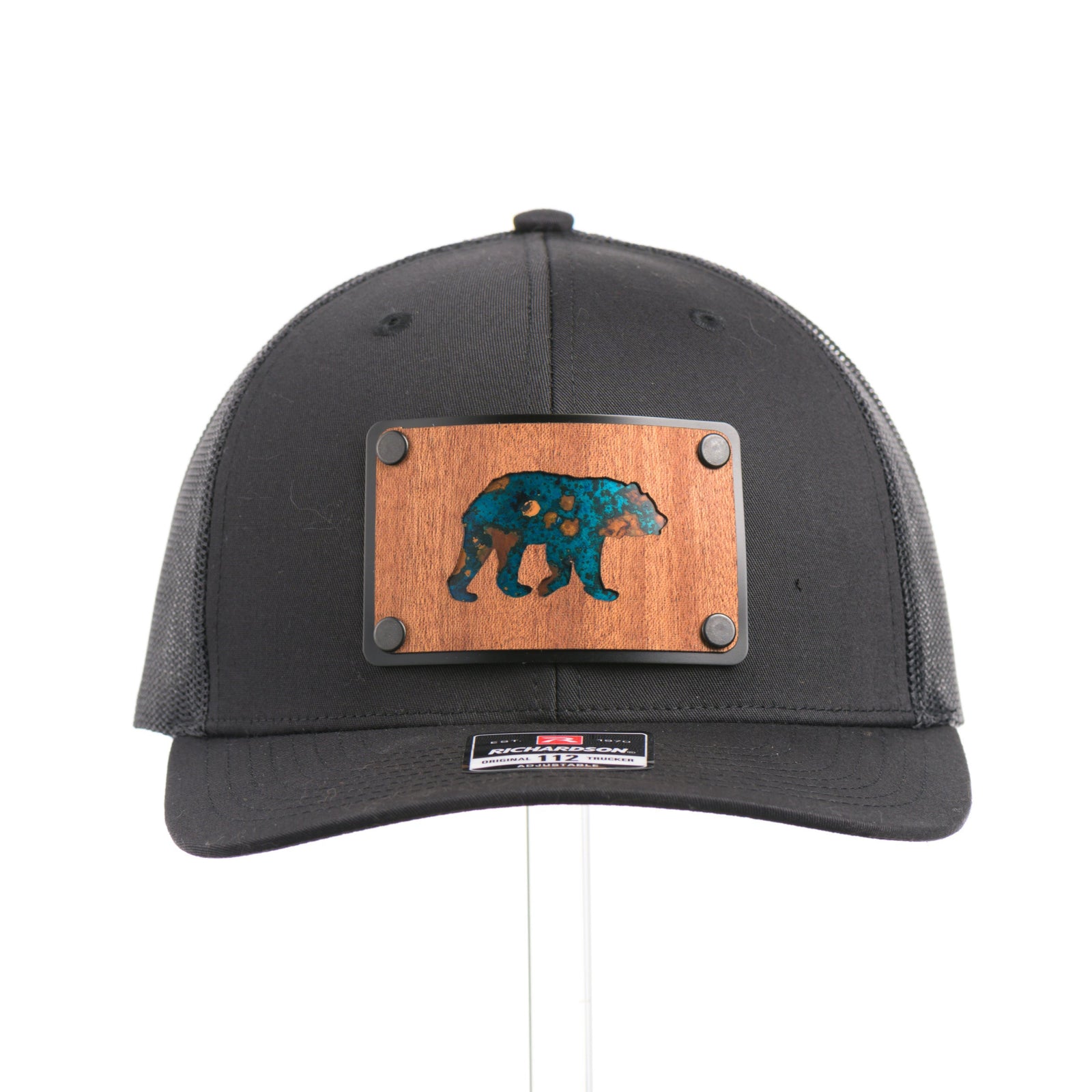 The front of a Black Grizzly Bear Teal Copper & Mahogany Wood Plate Patch Trucker Hat, featuring a rectangular real mahogany wood veneer patch with cut-out bear silhouette and teal copper inlay, laminated to a black anodized metal plate, and riveted to a black Richardson 112 trucker hat—handmade in Montana, USA by Last Best Supply Co.