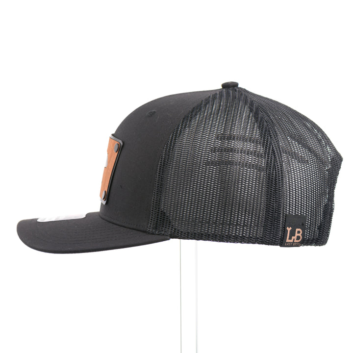 The side of a Black Grizzly Bear Teal Copper & Mahogany Wood Plate Patch Trucker Hat, featuring a rectangular real mahogany wood veneer patch with cut-out bear silhouette and teal copper inlay, laminated to a black anodized metal plate, and riveted to a black Richardson 112 trucker hat—handmade in Montana, USA by Last Best Supply Co. 