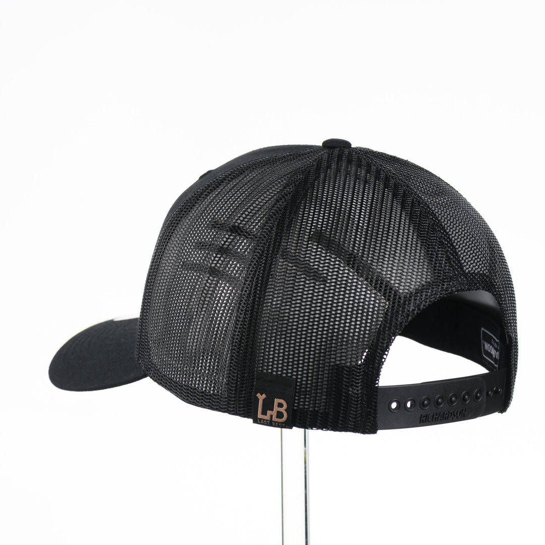Back of a Black Trucker Hat with Flexwood Patch Centennial Coin, featuring a hexagonal Flexwood veneer patch reverse etched with our iconic MT steer skull logo in a circle with the words "Montana 1889,” and "Big Sky Country," securely attached to a solid black trucker hat—handmade in Montana, USA by Last Best Supply Co.