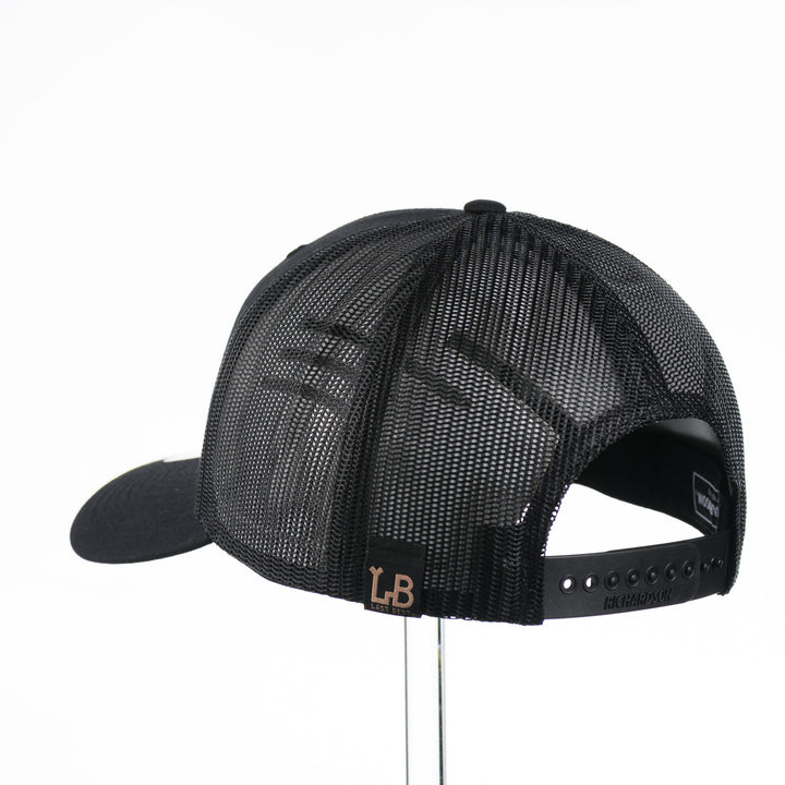 Back of a Black Trucker Hat with Flexwood Patch Centennial Coin, featuring a hexagonal Flexwood veneer patch reverse etched with our iconic MT steer skull logo in a circle with the words "Montana 1889,” and "Big Sky Country," securely attached to a solid black trucker hat—handmade in Montana, USA by Last Best Supply Co.