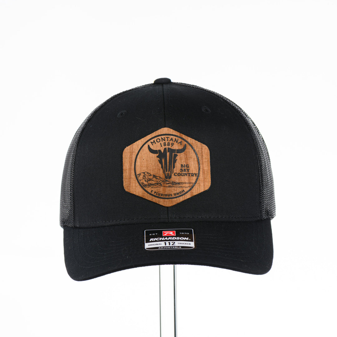 Front of a Black Trucker Hat with Flexwood Patch Centennial Coin, featuring a hexagonal Flexwood veneer patch reverse etched with our iconic MT steer skull logo in a circle with the words "Montana 1889,” and "Big Sky Country," securely attached to a solid black trucker hat—handmade in Montana, USA by Last Best Supply Co.