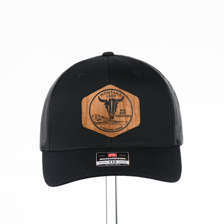Front of a Black Trucker Hat with Flexwood Patch Centennial Coin, featuring a hexagonal Flexwood veneer patch reverse etched with our iconic MT steer skull logo in a circle with the words "Montana 1889,” and "Big Sky Country," securely attached to a solid black trucker hat—handmade in Montana, USA by Last Best Supply Co.
