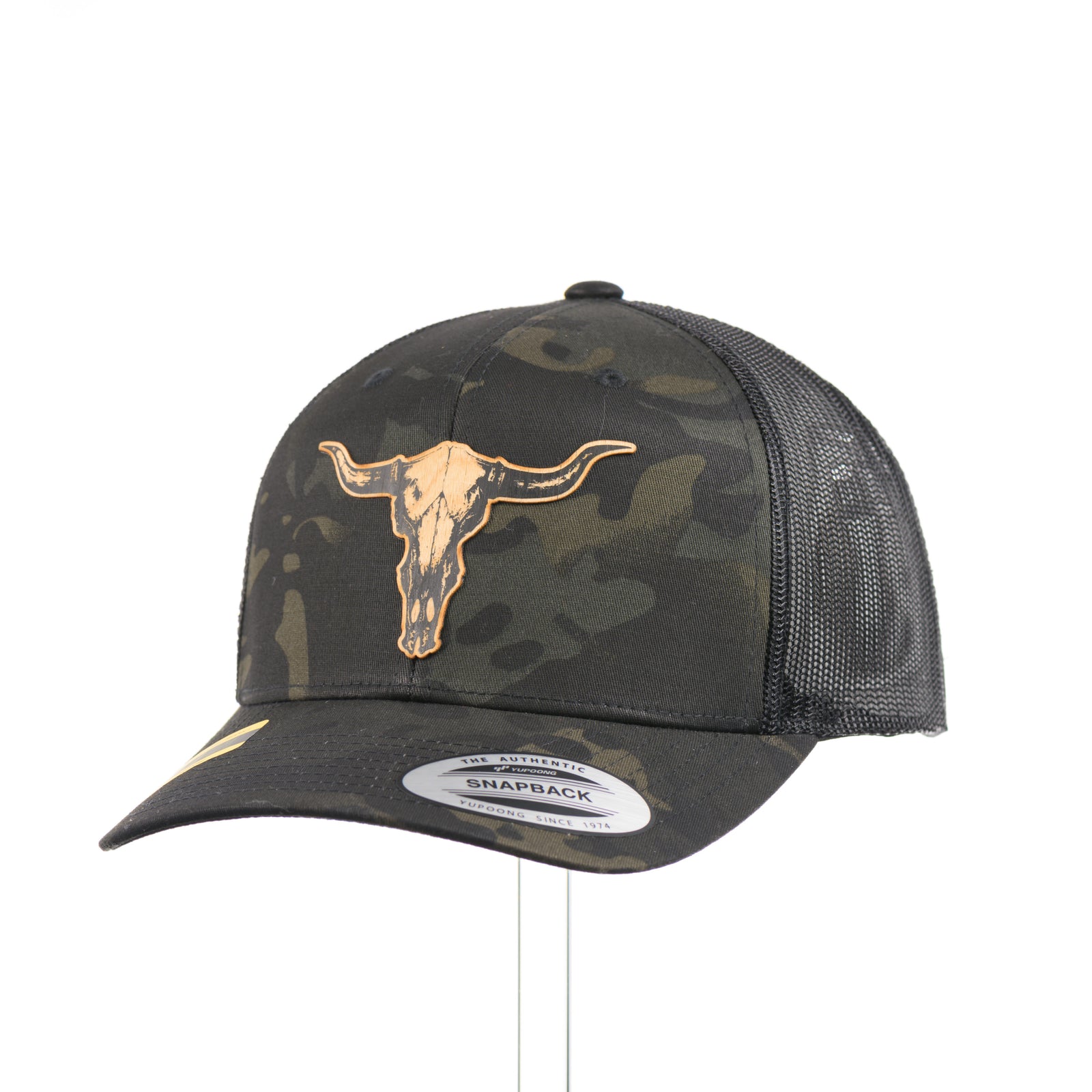 A dark multicam trucker hat with a flexwood steer skull patch at a quarter turn, handmade in Montana USA by Last Best Supply Co.