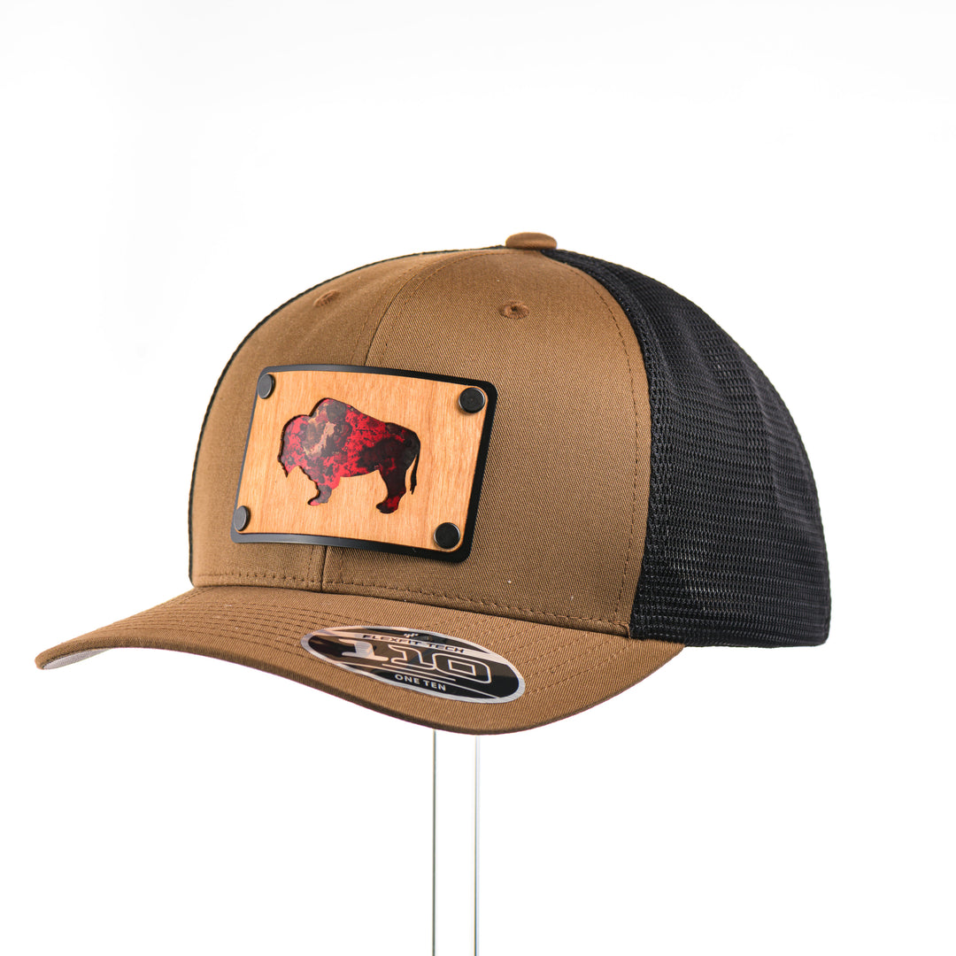 3/4 view of a Cherry Wood & Red Copper Bison Coyote Brown & Black Trucker Hat, featuring a rectangular real cherry wood veneer patch with cut-out bison silhouette inlayed with red copper, laminated to a black anodized aluminum plate, and riveted to a coyote brown and black mesh Flexfit 110M trucker hat—handmade in Montana, USA by Last Best Supply Co.