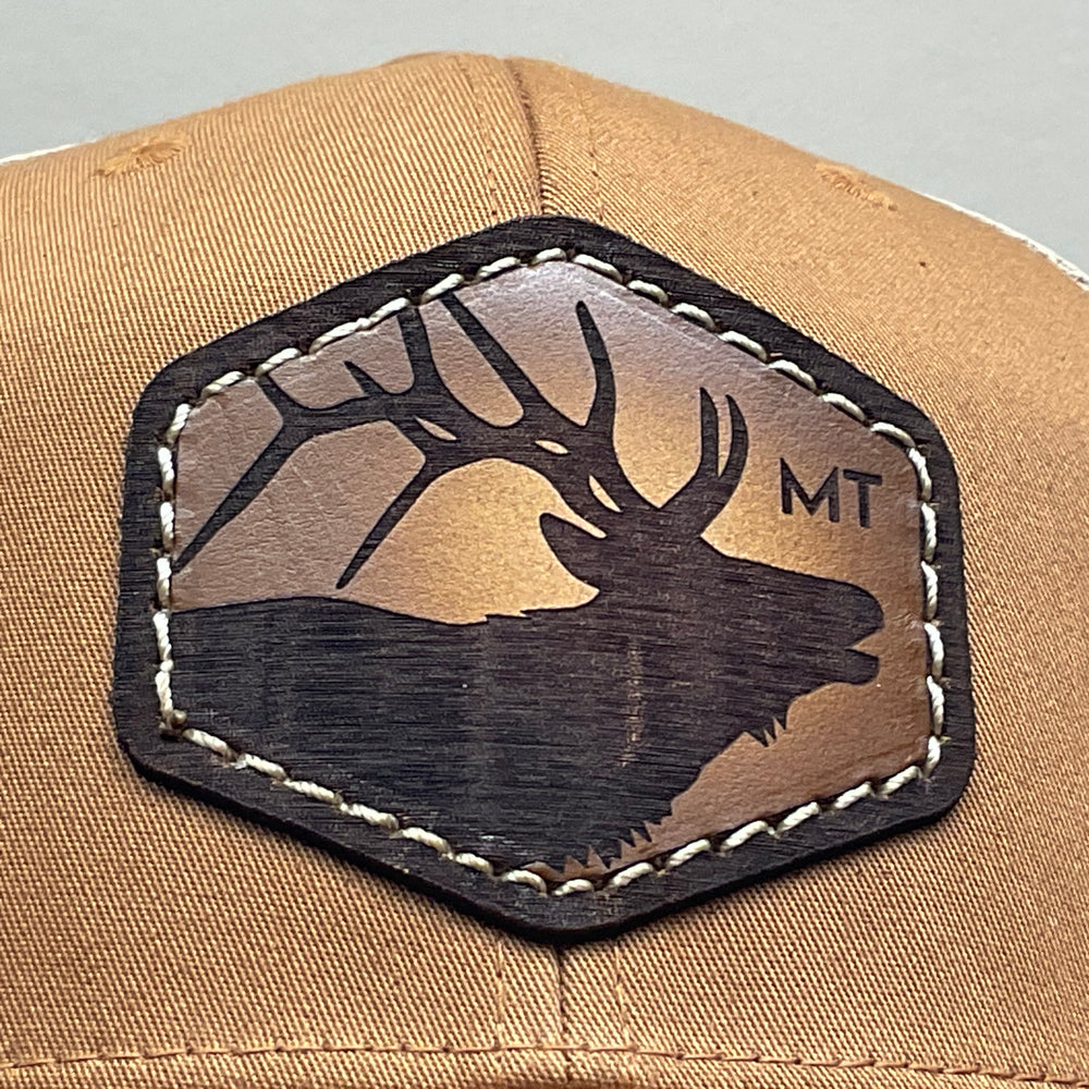Patch detail of an Elk Hand Dyed Leather Patch Flexfit Hat, featuring a hexagonal hand-dyed leather patch etched with the silhouette of an elk and the letters “MT”, securely sewn to the structured front of a camel and cream colored Flexfit trucker hat—handmade in Montana, USA by Last Best Supply Co.