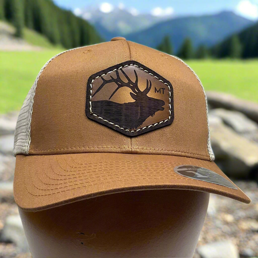 Front of an Elk Hand Dyed Leather Patch Flexfit Hat, featuring a hexagonal hand-dyed leather patch etched with the silhouette of an elk and the letters “MT”, securely sewn to the structured front of a camel and cream colored Flexfit trucker hat—handmade in Montana, USA by Last Best Supply Co.