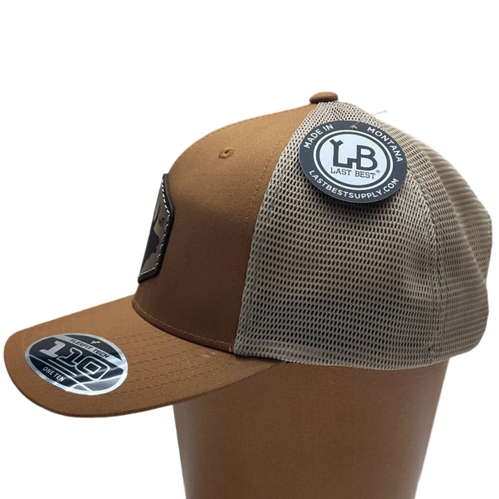 Side view of an Elk Hand Dyed Leather Patch Flexfit Hat, featuring a hexagonal hand-dyed leather patch etched with the silhouette of an elk and the letters “MT”, securely sewn to the structured front of a camel and cream colored Flexfit trucker hat—handmade in Montana, USA by Last Best Supply Co.