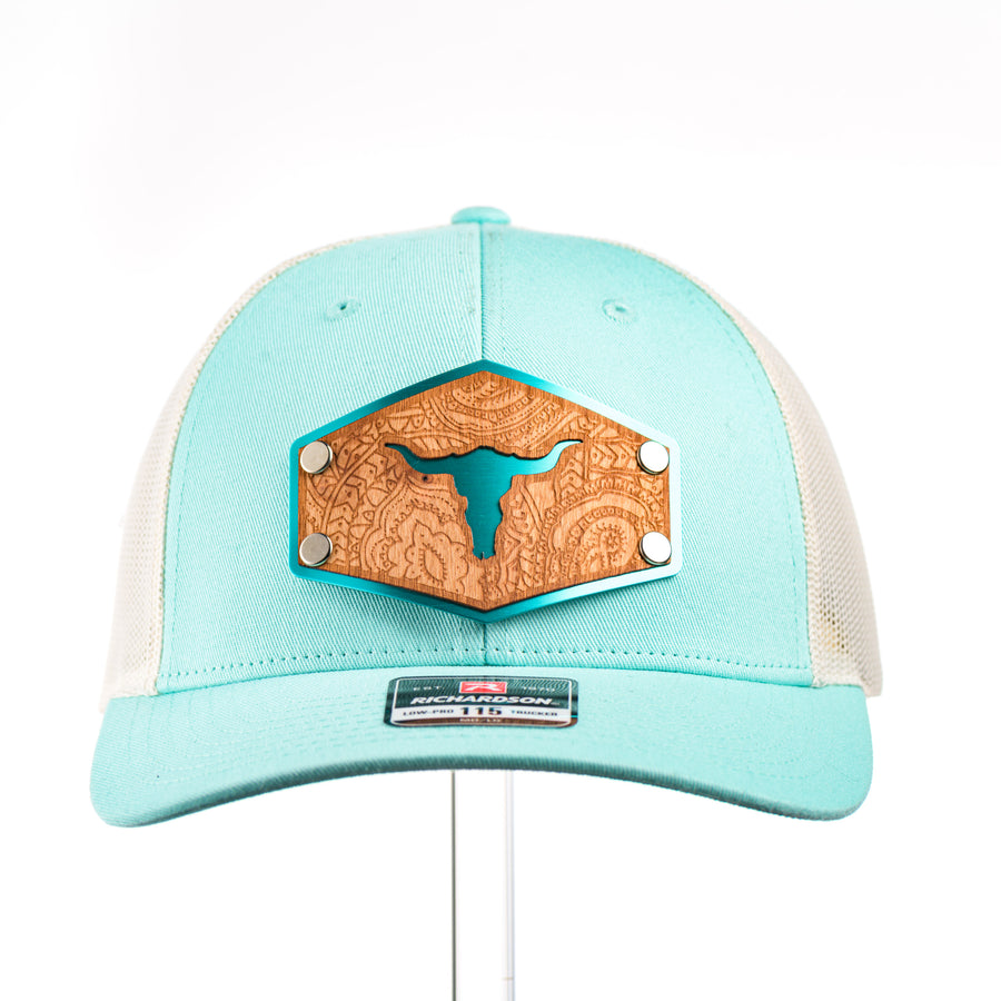 The front of a teal and cream mesh low profile Richardson trucker hat with an etched cherry wood cut out steer skull and paisley design on a hexagon metal patch.